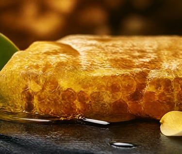 Golden raw honeycomb with dripping honey, green leaf and flower petals on a dark slate surface.