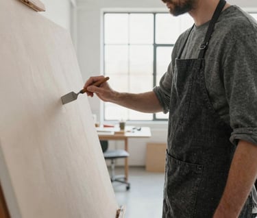 An over-the-shoulder shot of an artist in a minimalist North American studio. The person wears a charcoal linen apron and works on a large canvas using a palette knife. The room is filled with bright, diffused natural light from large industrial windows.