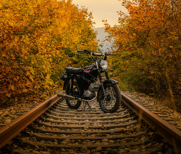 Vintage black motorcycle parked on railroad tracks surrounded by golden autumn forest foliage.