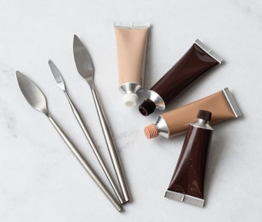 A minimalist arrangement of art tools including several stainless steel palette knives and tubes of acrylic paint in soft tan and dark brown on a white marble surface. North American / Western minimalist aesthetic.