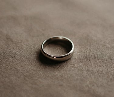 A minimalist detail shot of a wedding ring resting on a textured warm taupe fabric. The lighting is soft side-light, creating gentle shadows. The palette consists of dark charcoal, warm taupe, and soft off-white, embodying a serene and sophisticated artistry.