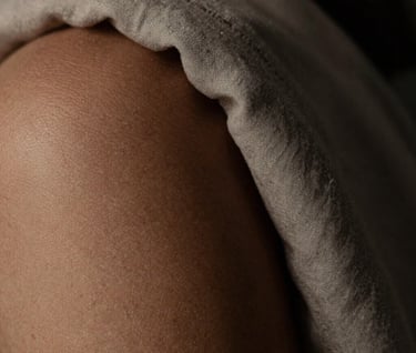 Macro photography showing the delicate texture of skin and a piece of muted sand-colored fabric. The lighting is low-key, creating deep dark charcoal shadows and a sense of calm artistry.