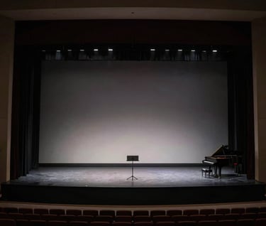A wide, cinematic shot of a minimalist music performance stage in a North American city. The space is dark and elegant with charcoal accents and soft gray backlighting that creates a sense of scale and professional prestige.
