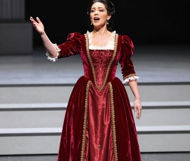 A professional production photograph of a mezzo-soprano in an elegant, red velvet period costume performing on a modern stage. The lighting is dramatic and focused, highlighting the artist's expression. North American / US opera house setting.