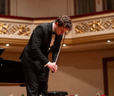 A medium shot of a classical musician during a curtain call in a sophisticated US concert hall, bowing gracefully under warm stage lights with red floral bouquets at her feet.
