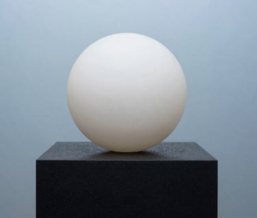 A minimalist art object featuring a smooth off-white sphere resting on a textured rich black pedestal. The background is a clean light steel blue. Sophisticated composition, sharp focus, capturing an avant-garde aesthetic.