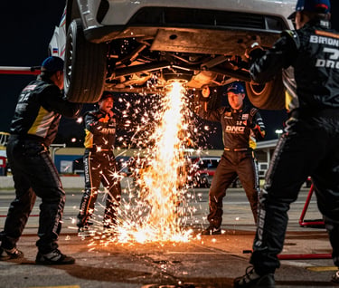 A dynamic shot of a pit crew in North American / US racing gear during a nighttime pit stop, sparks flying from the car underbody, captured with high-speed photography to freeze the frantic motion and dramatic light.