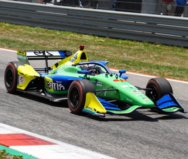 A high-speed action shot of a vibrant IndyCar navigating a street circuit in the North American / US sun. Intense motion blur on the wheels and track surface, emphasizing the raw energy and speed. Professional photography style with a focus on color and precision.