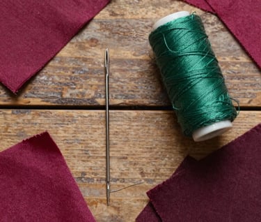 A flat lay photograph of sewing tools—a vintage needle, forest green thread, and dark magenta fabric scraps—arranged on a rustic wooden table in a whimsical workshop, Western / Global.