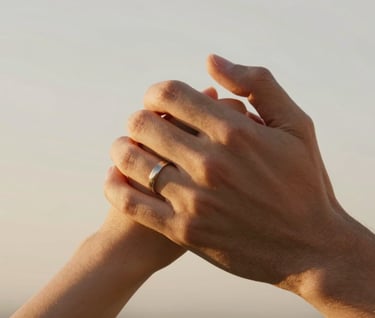 A close-up of two hands intertwined, wedding rings catching the warm golden light, skin tones looking natural and authentic against a soft off-white background.