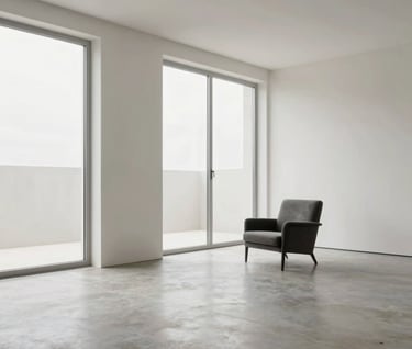 A wide interior photograph of a minimalist residential living room. It features floor-to-ceiling windows, polished concrete floors in light grey, and a single charcoal-colored armchair. The walls are an elegant off-white (#FBF9F6). The lighting is natural and serene, showcasing the volume of space. Style: Professional, high-end photography.