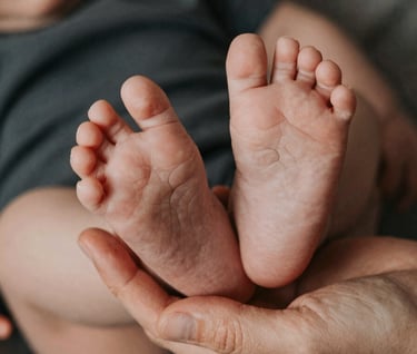 Details of a baby's tiny feet being held by a parent's hand. Anthracite and terracotta color accents in the clothing. Soft cinematic lighting, intimate and emotional atmosphere, European / French style.
