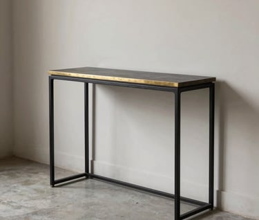 An elegant, slim-profile console table made of blackened steel with a polished brass inlay. The table is placed in a minimalist hallway with soft off-white walls and concrete flooring.