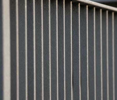 Detail shot of rhythmic vertical steel bars in a slate grey finish, forming a clean-lined railing for a mezzanine. The lighting is soft and natural, emphasizing the 'warm minimalism' aesthetic.