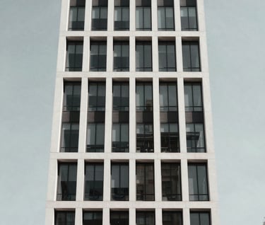 A low-angle shot of a minimalist modern building facade in New York City. The structure features clean white lines and glass against a clear, pale gray sky. The composition is centered and bold, reflecting a sophisticated and aspirational urban vibe.