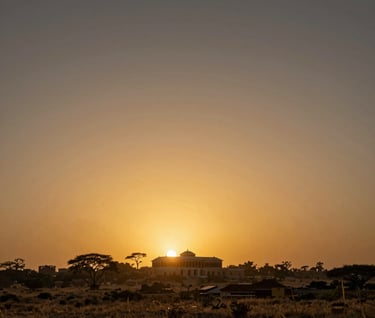 Wide-angle landscape shot of a lush African horizon at sunset, silhouetting a traditional palace structure. The sky is a mix of #A88B57 gold and deep dark tones, establishing an epic scale for the production.