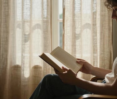 Candid photography of a person reading a book in a sunlit room, linen curtains, warm sand tones, authentic European / Portuguese home interior.