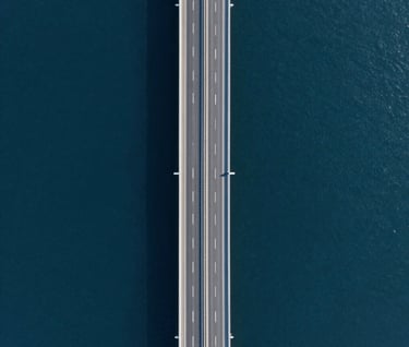 An aerial drone shot looking straight down at a modern coastal highway. The water is deep navy #1B263B and the road is sharp grey #415A77. The composition is symmetrical and high-precision, showcasing high-fidelity drone footage quality.