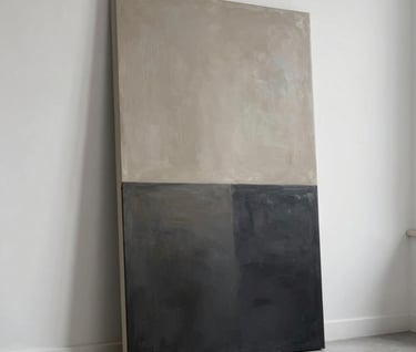 A vertical abstract painting leaning against a minimalist white wall in a sunlit artist's studio. The artwork displays a gradient from deep charcoal at the bottom to a hazy muted taupe at the top. The overall mood is sophisticated and peaceful.