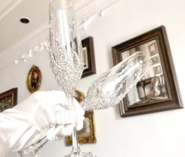 A gloved hand holds luxury crystal champagne flutes decorated with silver glitter and rhinestones.