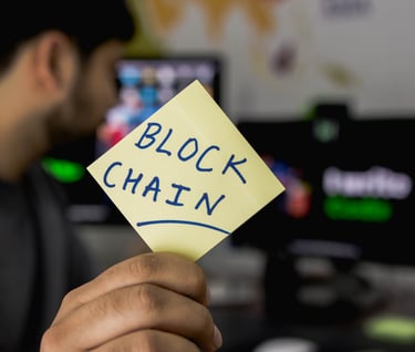 A man facing away from the camera holding up and post it note that says, "Blockchain".