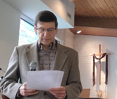 An elderly man with dark hair wearing a gray jacket and checkered shirt, holding a microphone and document in front of cross