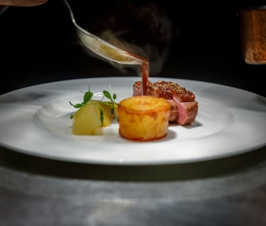 Chef pouring red wine jus over gourmet pan-seared duck breast with potato fondant and garnish.