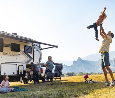 A multi-generational family enjoys an RV camping trip in the mountains while a father lifts his child.