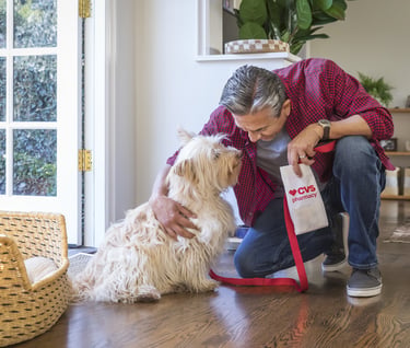 A man at home petting his fluffy dog while holding a CVS Pharmacy prescription bag for pet medication.