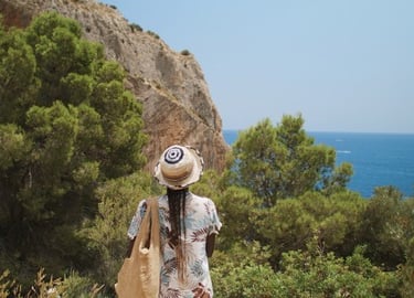 A woman in a sun hat looking at the blue Mediterranean Sea from a rocky cliff with green pine trees.