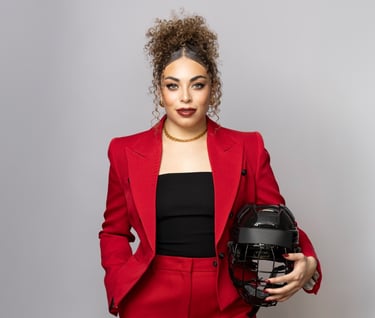 A woman in a red power suit posing with a black hockey helmet against a grey background.