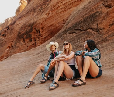 Friends sitting on red rock canyon formations wearing casual summer hiking clothes and outdoor sandals.
