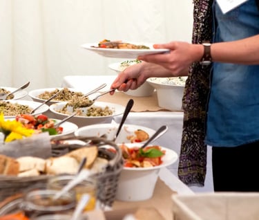 A person serving fresh healthy food from a catered buffet line featuring quinoa salad and bread.