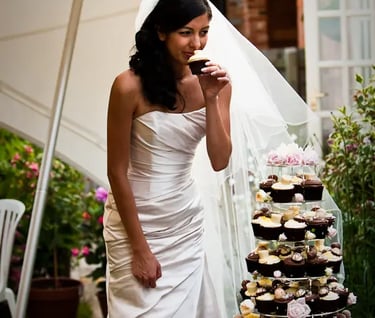 A bride in a white wedding gown eating a chocolate cupcake by a tiered cupcake tower.