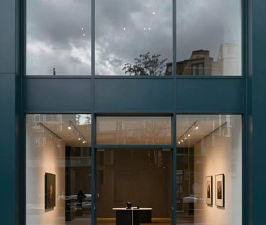 A portrait-oriented photography shot of a sleek, modern Northern European / Welsh / British gallery entrance with a large glass window reflecting a cloudy sky, showcasing a minimalist interior with deep charcoal teal accents.