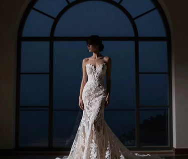 Artistic silhouette of a bride wearing a high-fashion lace gown, standing against a large arched window with a dark midnight blue background. Minimalist composition, emphasizing the delicate details of the dress and the elegance of the pose.
