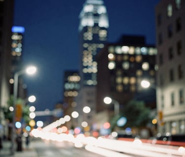 Artistic photography of motion-blurred lights in a North American / US cityscape at night. The dominant colors are deep navy blue and sharp off-white, creating a sense of energy and transformation. The composition is abstract, focusing on the atmosphere of a bustling metropolis.
