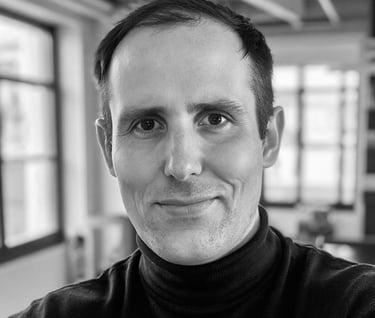 Black and white professional headshot of a smiling man wearing a dark turtleneck sweater in an office.