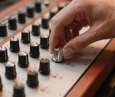 Detailed close-up of a hand adjusting a silver knob on a boutique analog synthesizer. The scene is bathed in warm terracotta lighting, highlighting the premium craftsmanship of the musical gear.