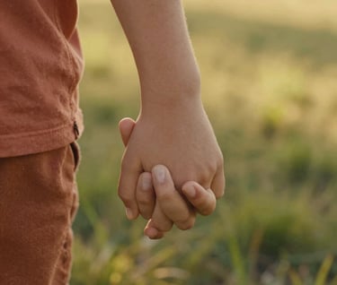 A close-up, artistic shot of a child's hand holding a parent's hand against a background of sun-dappled grass. The lighting is warm and golden. Soft focus, cinematic storytelling style, highlighting textures of skin and terracotta-colored fabric.