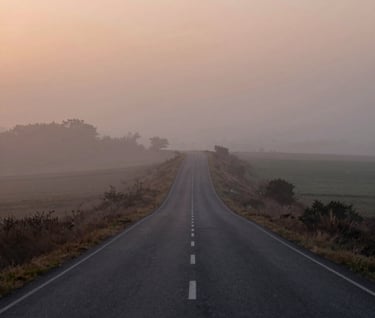 A wide-angle shot of a winding country road disappearing into a soft mist at sunset. The lighting is ethereal and hazy, using #F5F0E1 for the sky and #262626 for the road. The style is timeless and cinematic.