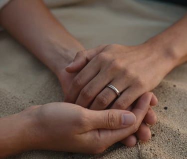 A detailed close-up of two hands holding, one with a wedding band, resting on a soft sand colored blanket. Warm, cinematic lighting with terracotta highlights, shot in a North American / US outdoor setting.
