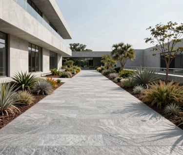 Exterior shot of a walkway made of light silver gray stone. The path is flanked by low-impact local vegetation and clean architectural lines of the main building under a clear sky.