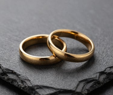 Macro photography of two gold wedding bands resting on a dark slate surface. Sophisticated side-lighting, shallow depth of field, elegant and minimalist composition, Southern European / Spanish luxury context.