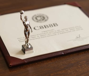 A close-up artistic shot of a silver vocal trophy and a leather-bound certificate resting on a mahogany surface. The lighting is soft and warm, incorporating #C8B8B8 and #8D6A6A tones to evoke a sense of prestigious history.