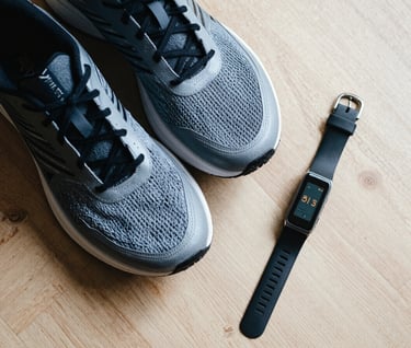An aesthetic overhead shot of sleek running shoes and a fitness tracker placed neatly on a light wood floor. The lighting is soft and natural, emphasizing a minimalist and personal lifestyle vibe.