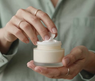 Close-up of hands with minimalist rings opening a Warm Off-White cream jar. The composition is clean and centered, featuring Muted Sage textiles in the soft-focus background.