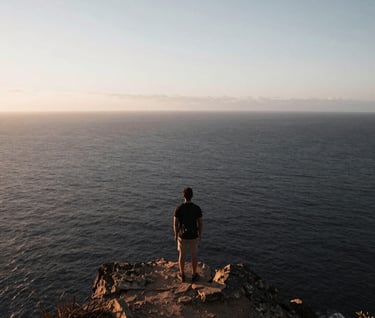 A wide-angle shot of a lone traveler standing on a cliff overlooking a deep charcoal ocean. The sky is a soft silver dawn color. Minimalist and inspiring travel photography.