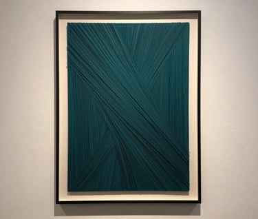 A vertical shot of a framed piece of generative art hanging on an off-white gallery wall. The art consists of millions of fine deep teal lines creating a sense of movement. The lighting is soft and contemplative.