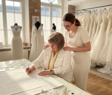 A bridal gown designer sketches a wedding dress pattern with an assistant in a sunlit bridal boutique.